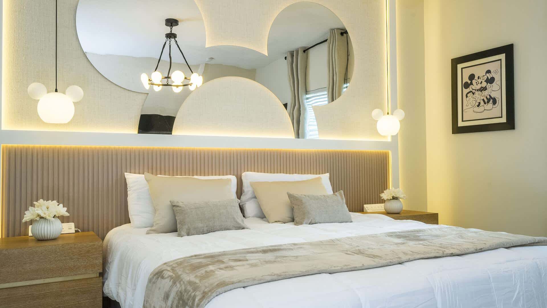 Ein Hotelzimmer mit einem großen Bett, einem Kopfteil mit integrierter Beleuchtung, einem gerahmten Bild von Mickey Mouse und einem einzigartigen Deckenstil.