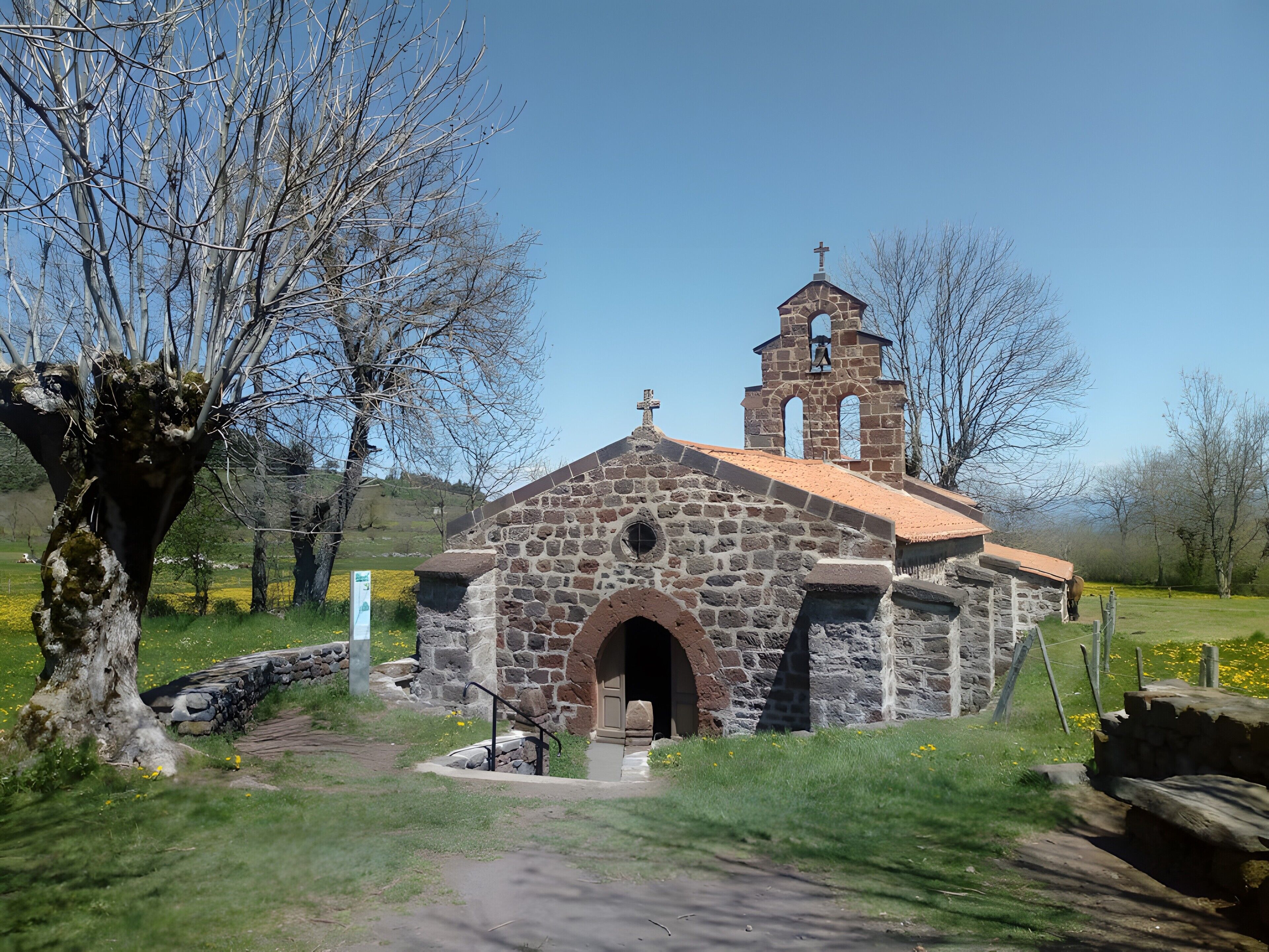 Une petite église en pierre, dotée d’un clocher et surmontée d’une croix, entourée d’arbres et bordée d’une pelouse.