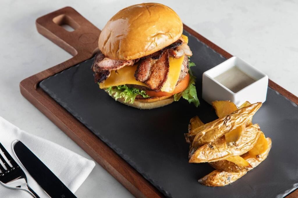 A burger with bacon, cheese, and lettuce on a bun, served with roasted potatoes and a small bowl of sauce on a black slate plate.