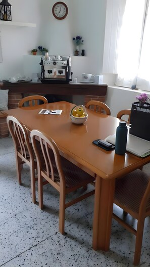 A dining area with a wooden table, chairs, a bowl of fruit, a water bottle, and a coffee machine.