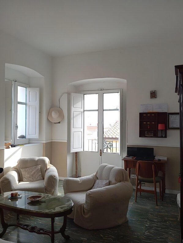 A living room with two sofas, a glass coffee table, a desk with a laptop, and a view through a large window.