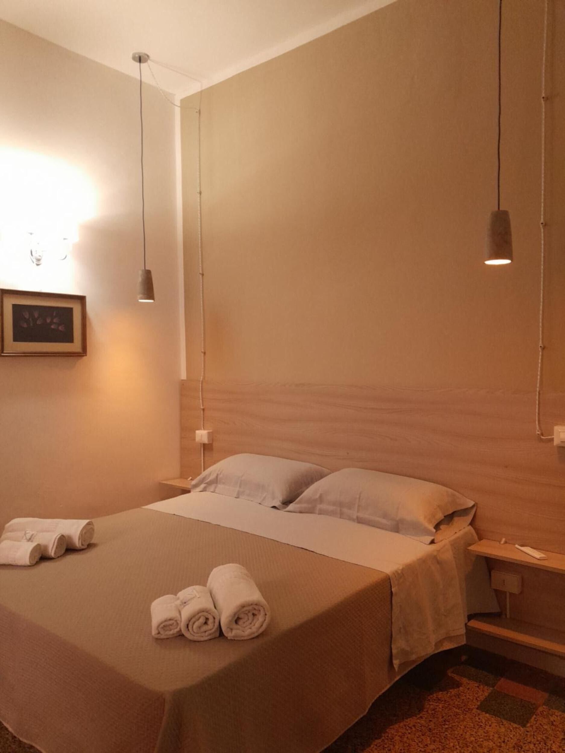 A hotel room with a bed, two pillows, a bedside table, a lamp, and rolled towels on the bed.