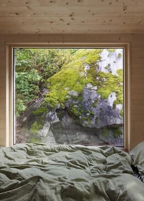 A room with a bed, a window with a view of a moss-covered rock and trees.