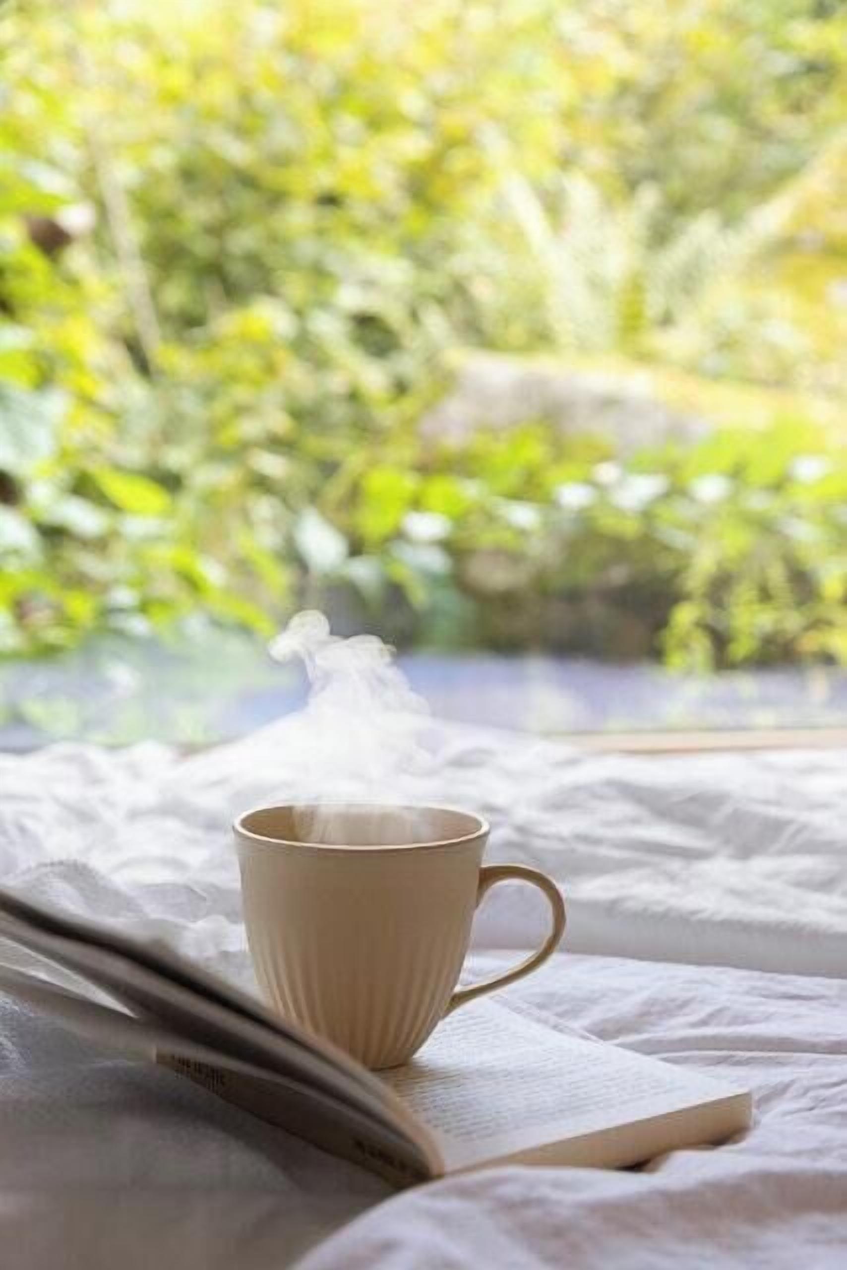 Una taza de bebida caliente sobre una cama, con un libro, cerca de una ventana con vistas a la vegetación.