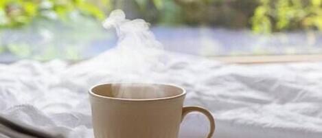 Una taza de bebida caliente sobre una cama, con un libro, cerca de una ventana con vistas a la vegetación.