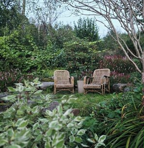 Zwei Korbstühle in einem Garten mit üppigem Grün und Steinen.