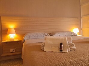 Ein Hotelzimmer mit einem Bett, zwei Nachttischlampen, einem gefalteten Handtuch und einer Flasche auf dem Bett.