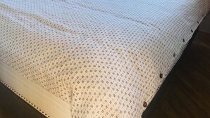 Una cama con una colcha estampada, una mesita de noche, una lámpara de pared y una ventana con vistas a árboles.