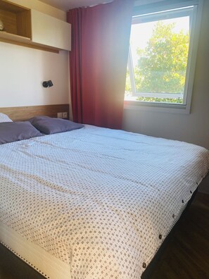 Ein Bett mit gemustertem Überwurf, ein Nachttisch, eine Wandlampe und ein Fenster mit Blick auf Bäume.