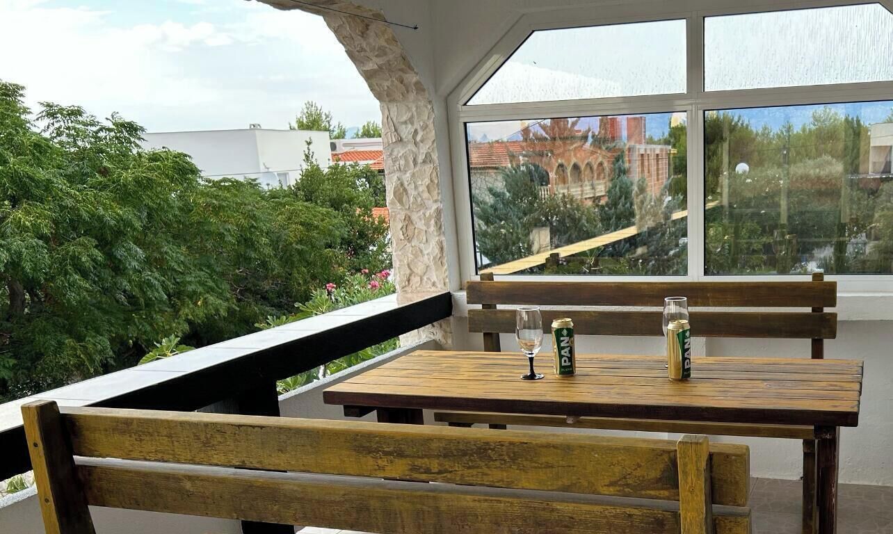 Ein Holztisch mit zwei Bänken, einem Glas und einer Bierdose auf einem Balkon mit Blick auf ein Gebäude und Grünflächen.