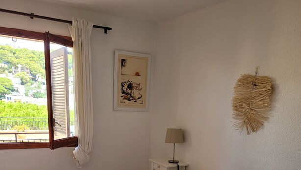 Ett sovrum med en säng, ett sängbord med en lampa, ett fönster med gardiner och en dekorativ detalj som hänger på väggen.