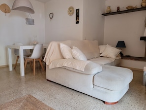 A living room with a sofa, a dining table with chairs, a lamp, and wall decorations. - Beach Apt: Huge 80m² Private Terrace, 2 Min Walk to Beach &  Free Parking (Tamariu)