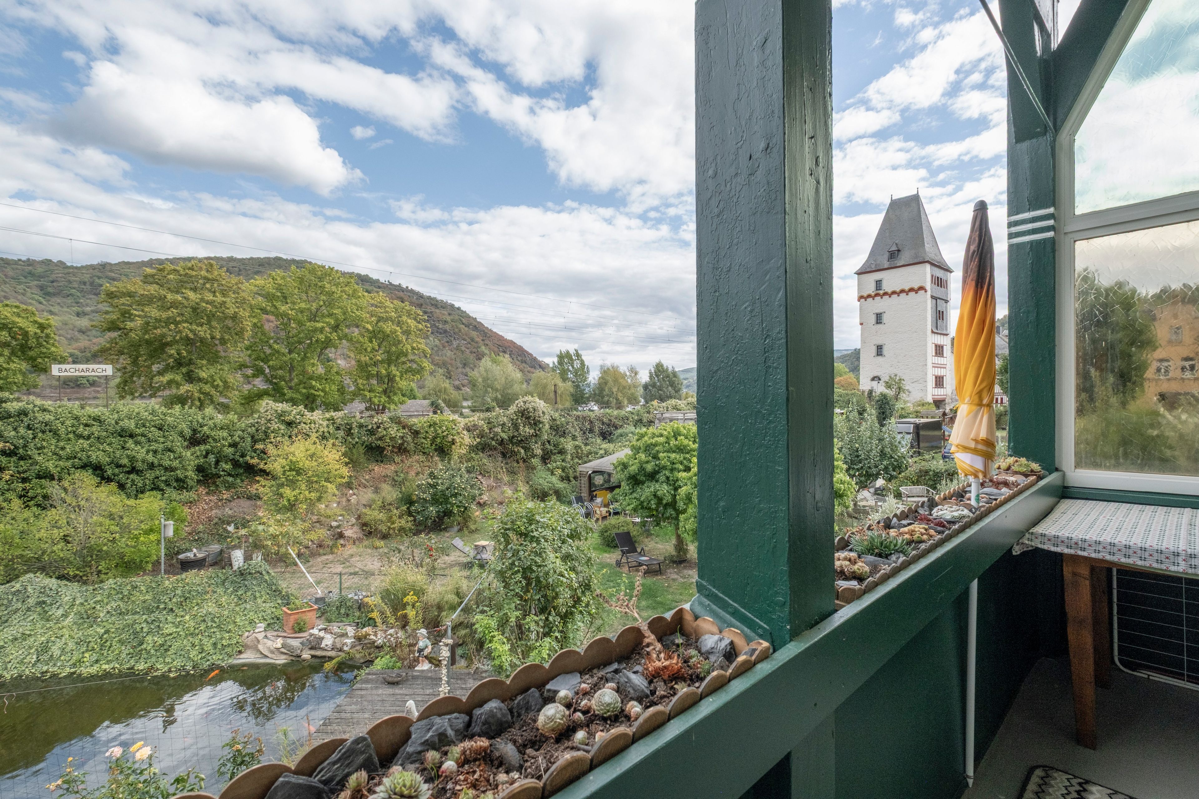 Une vue depuis une fenêtre donnant sur une rivière, un jardin avec des rochers et des plantes, et un bâtiment éloigné surmonté d’une tour.