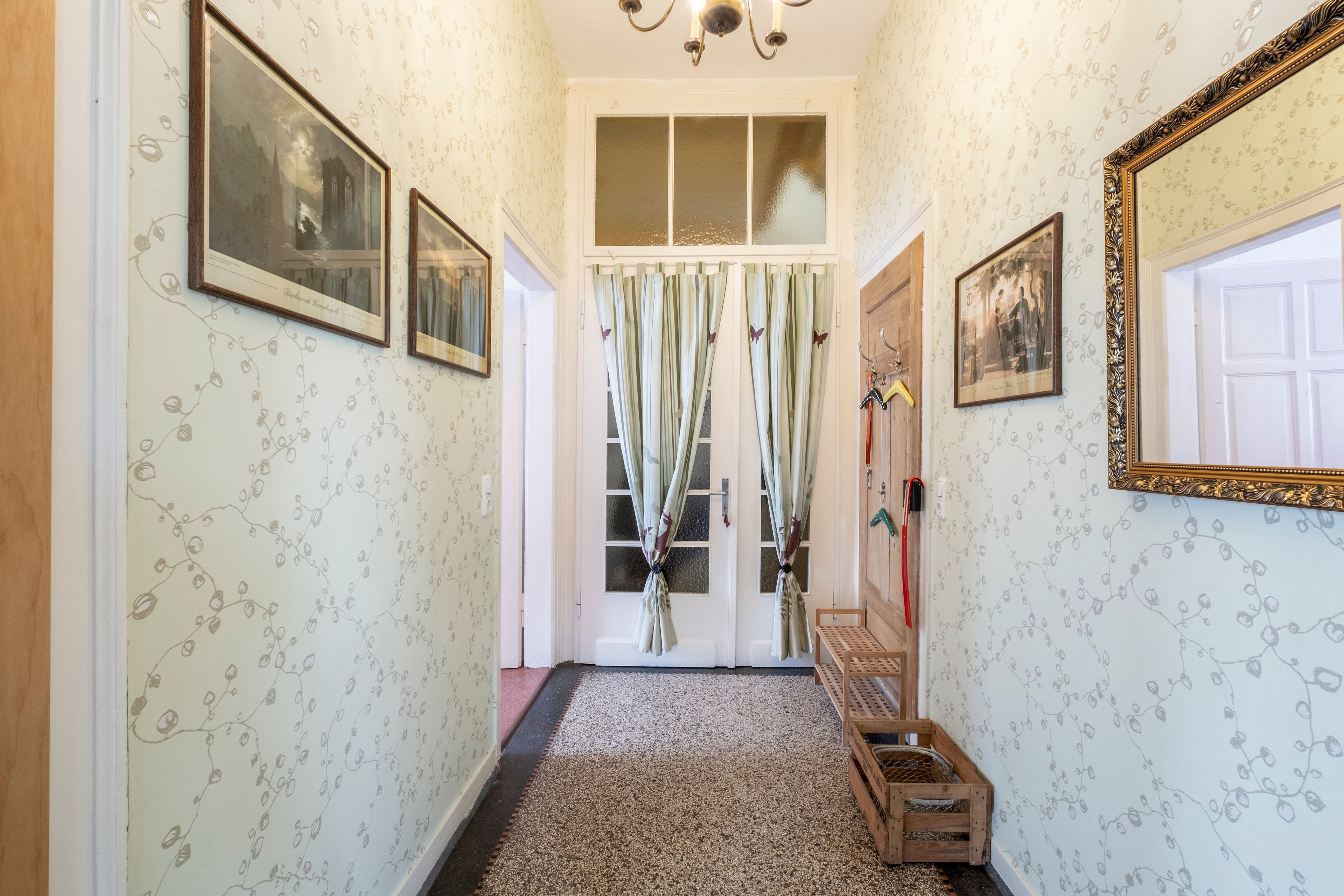 A hallway with patterned wallpaper, framed pictures on the walls, a wooden bench, and a door with curtains.