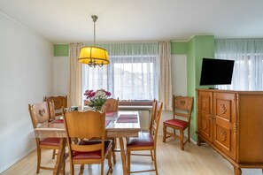 Un coin repas comprenant une table, des chaises, un lustre, un téléviseur et une fenêtre avec des rideaux.
