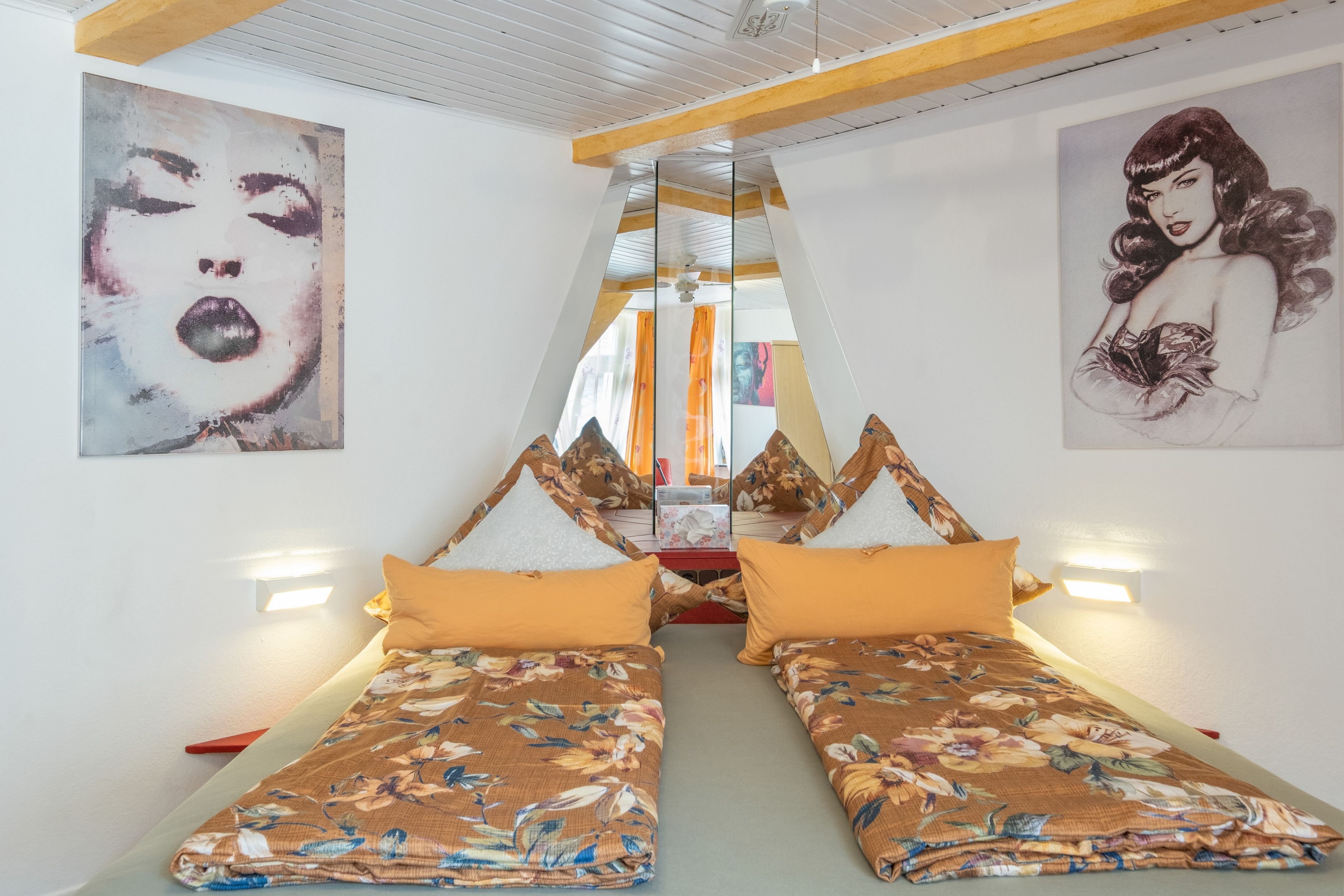 A room with two beds, each with floral bedding and orange pillows. There are wall-mounted lights on either side of the beds.