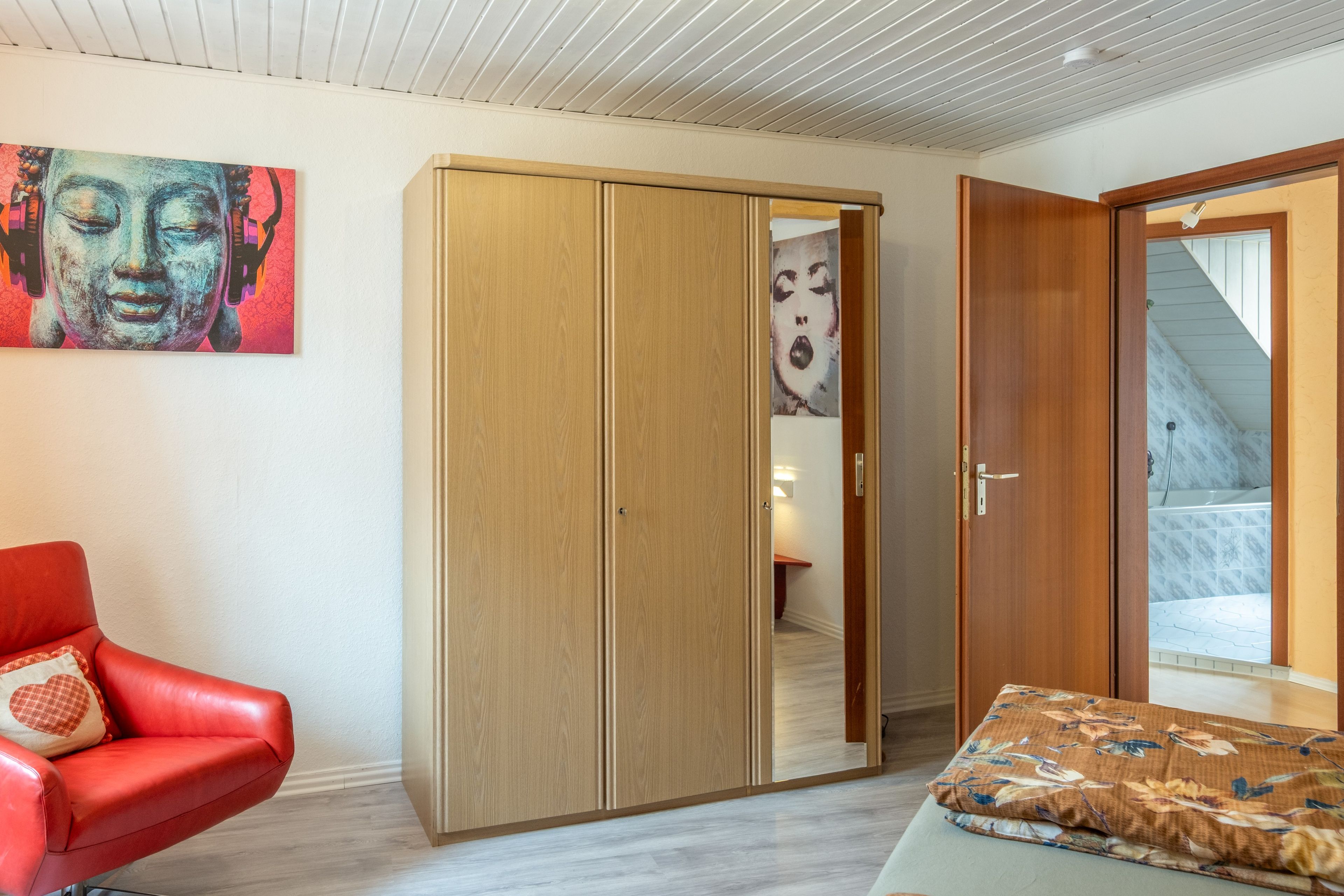 A room with a red leather chair, a wooden wardrobe, a bed with a floral pillow, and a painting on the wall.