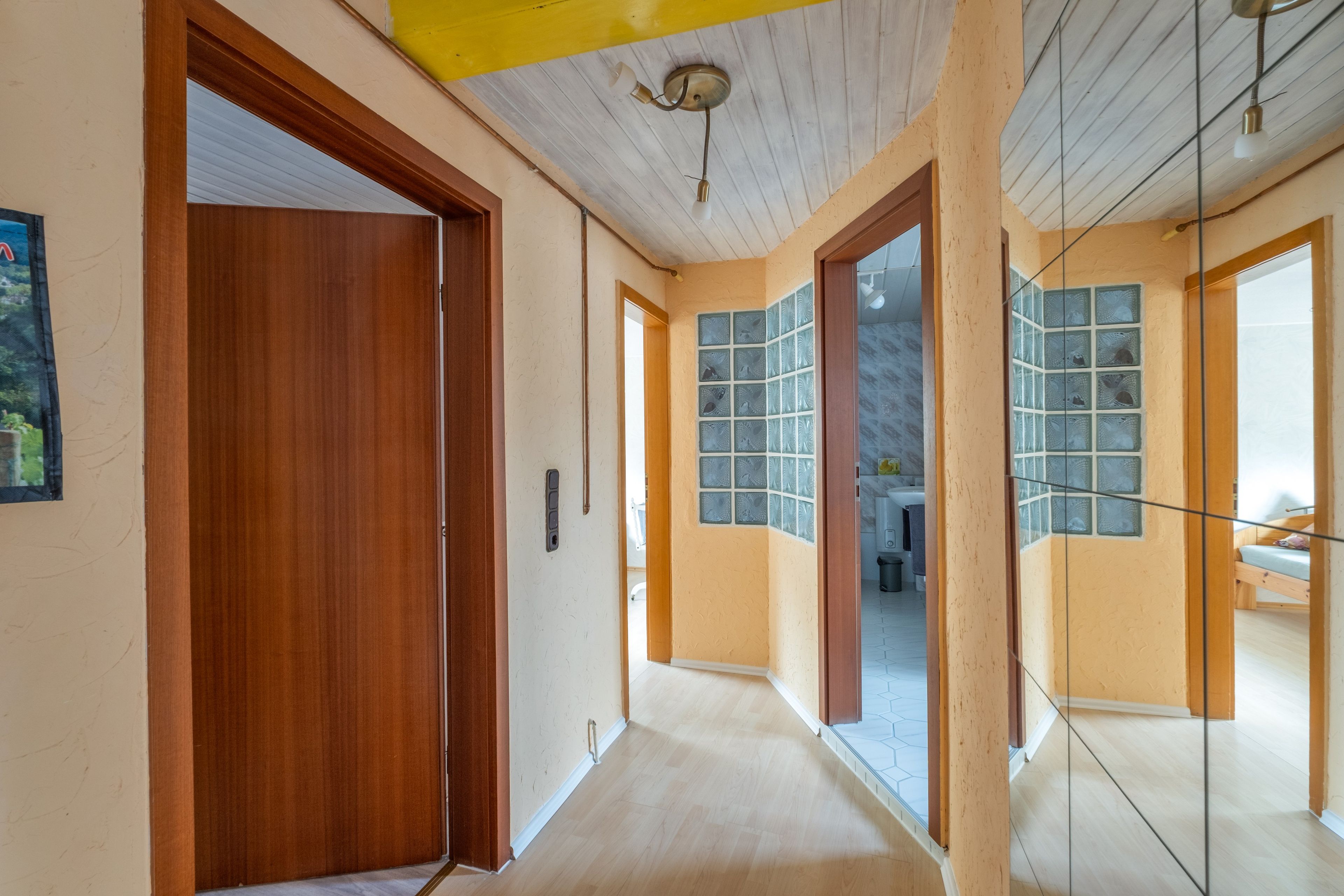 Un couloir avec des portes en bois, un ventilateur de plafond et une vue sur une pièce où se trouvent un lit et une fenêtre.