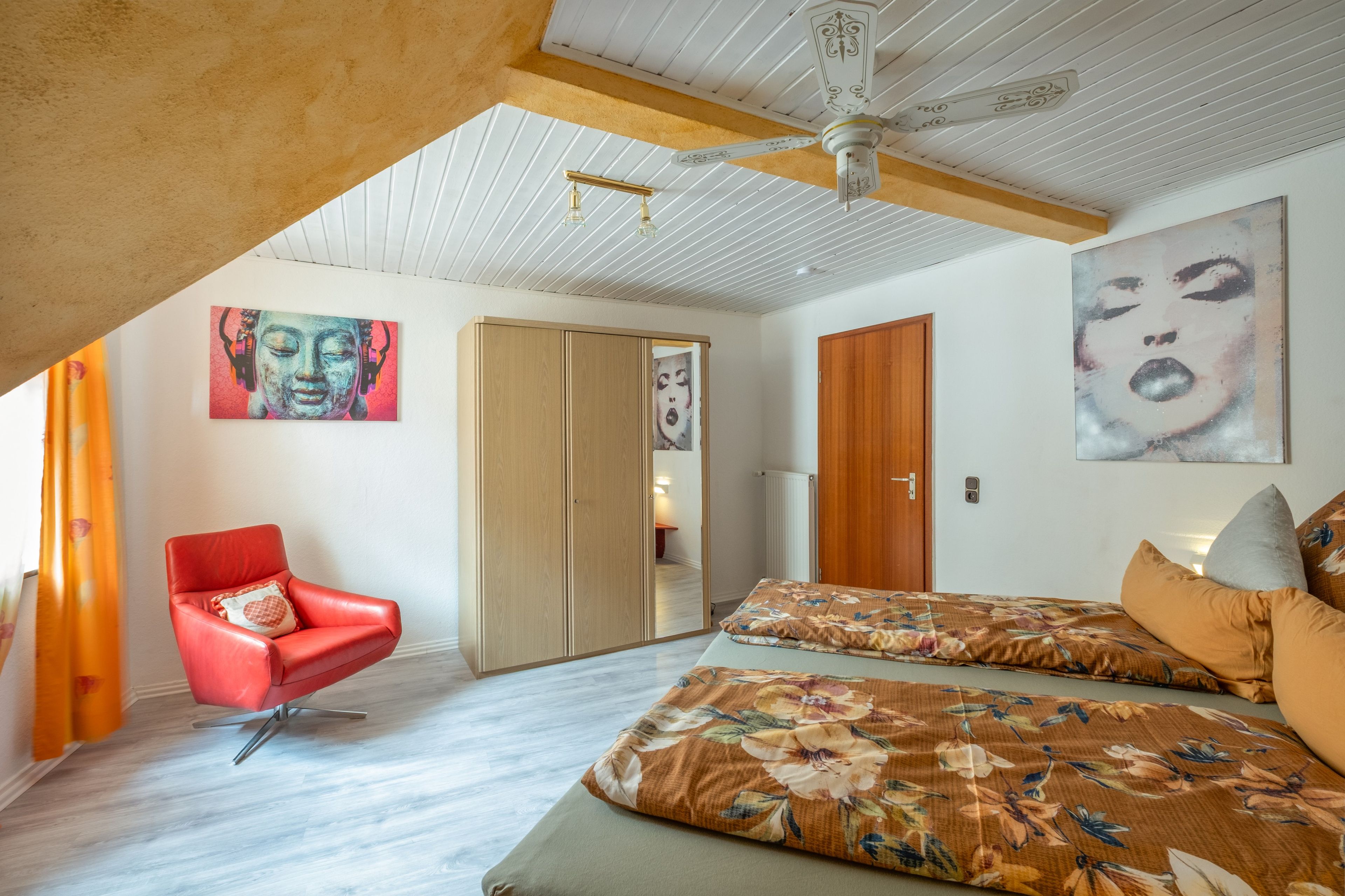 A bedroom with a sloped ceiling, a red chair, a bed with floral bedding, and two paintings on the walls.