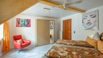 A bedroom with a sloped ceiling, a red chair, a bed with floral bedding, and two paintings on the walls.