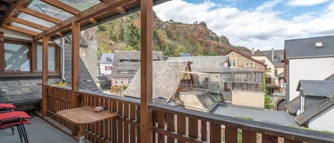 A wooden balcony with a table and chairs, overlooking a town with stone buildings and a hillside.