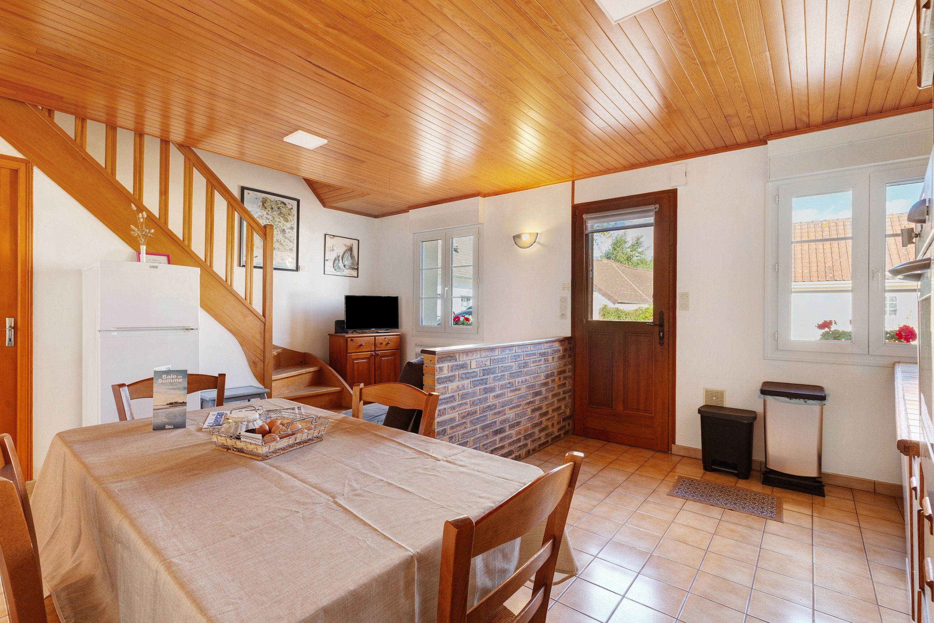Un coin repas avec une table en bois, des chaises, un téléviseur et un mur en briques.
