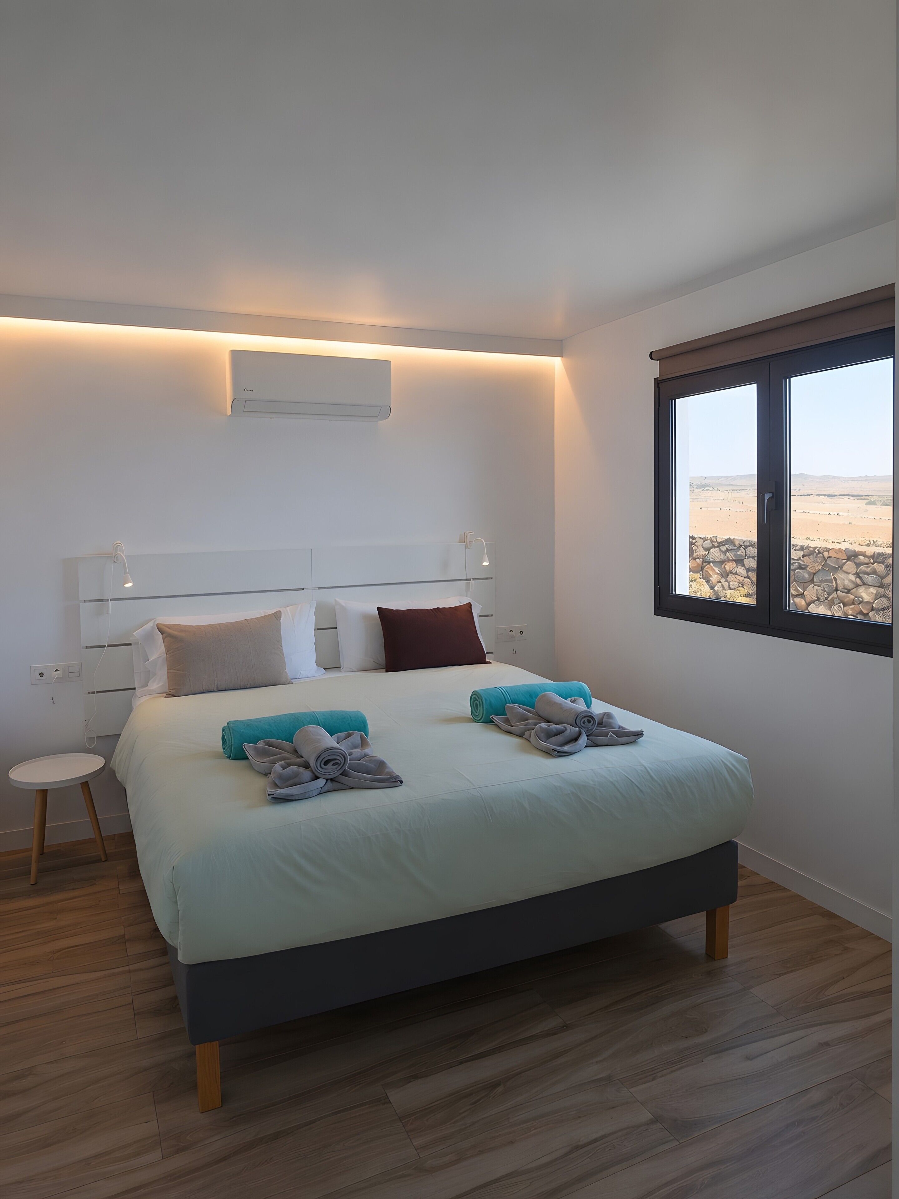 Ein modernes Schlafzimmer mit Bett, Nachttischen, einem Fenster mit Blick auf die Wüste und einer Wand-Klimaanlage.