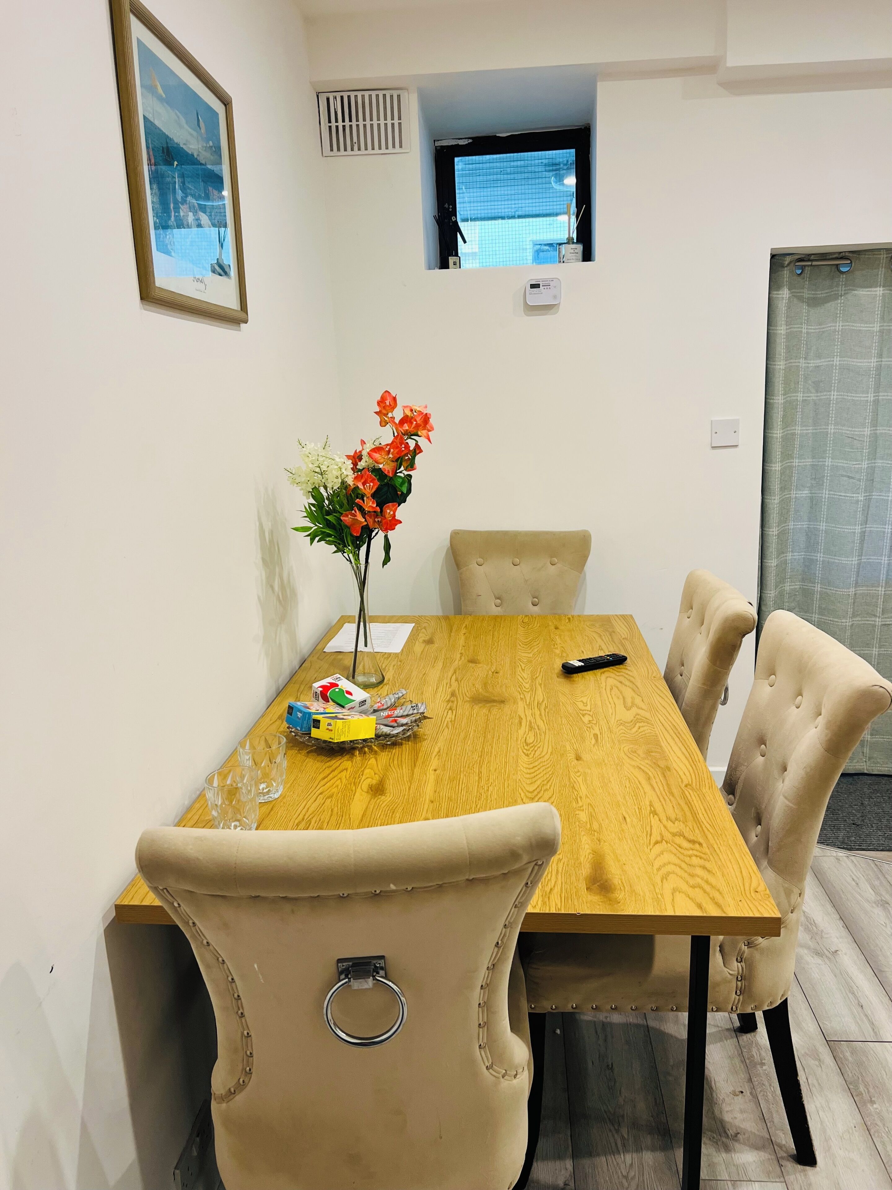 A dining area with a wooden table, beige upholstered chairs, a vase with flowers, and a television on the wall.