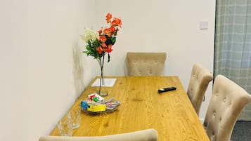 A dining area with a wooden table, beige upholstered chairs, a vase with flowers, and a television on the wall.