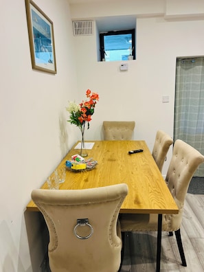 A dining area with a wooden table, beige upholstered chairs, a vase with flowers, and a television on the wall.