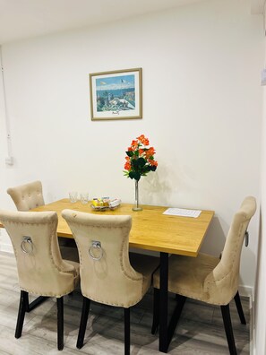 A dining area with a wooden table, four upholstered chairs, a vase with flowers, a framed picture on the wall, and a small table with a toy and glasses.