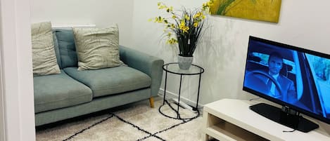 A living room with a grey sofa, a small side table, a flat-screen TV, a painting of red flowers, and a patterned rug.