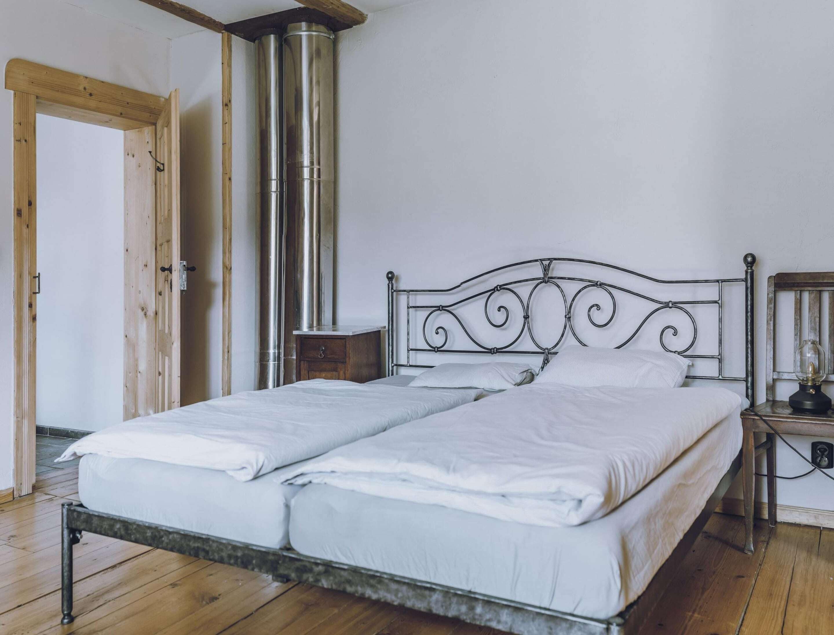 Una cama con estructura metálica y ropa de cama blanca, suelo de madera, puerta de madera y un calentador fijado a la pared.