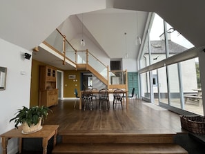 Ein geräumiges Interieur mit einer Holztreppe zu einem Schlafbereich, einem Esstisch mit Stühlen und großen Fenstern mit Ausblick.