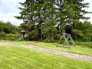 Ein Spielplatz mit Rutsche und Schaukeln in einer grünen, von Bäumen gesäumten Fläche.