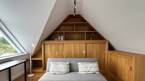 Ett sovrum på entresolplan med ett sluttande tak, en inbyggd bokhylla i trä, en grå soffa med randiga kuddar och ett skrivbord med en lampa.