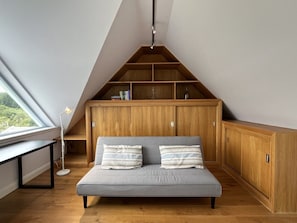 Ein Schlafzimmer im Loftsstil mit schrägem Dach, einer eingebauten Holzbücherregal, einem grauen Sofa mit gestreiften Kissen und einem Schreibtisch mit Lampe.
