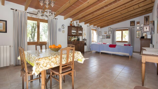 A spacious dining area with a wooden table and chairs, a chandelier, and a view of the outdoors through large windows. - Country Home “Servera” with Pool, Wi-Fi, Terrace & Garden (Santanyí)