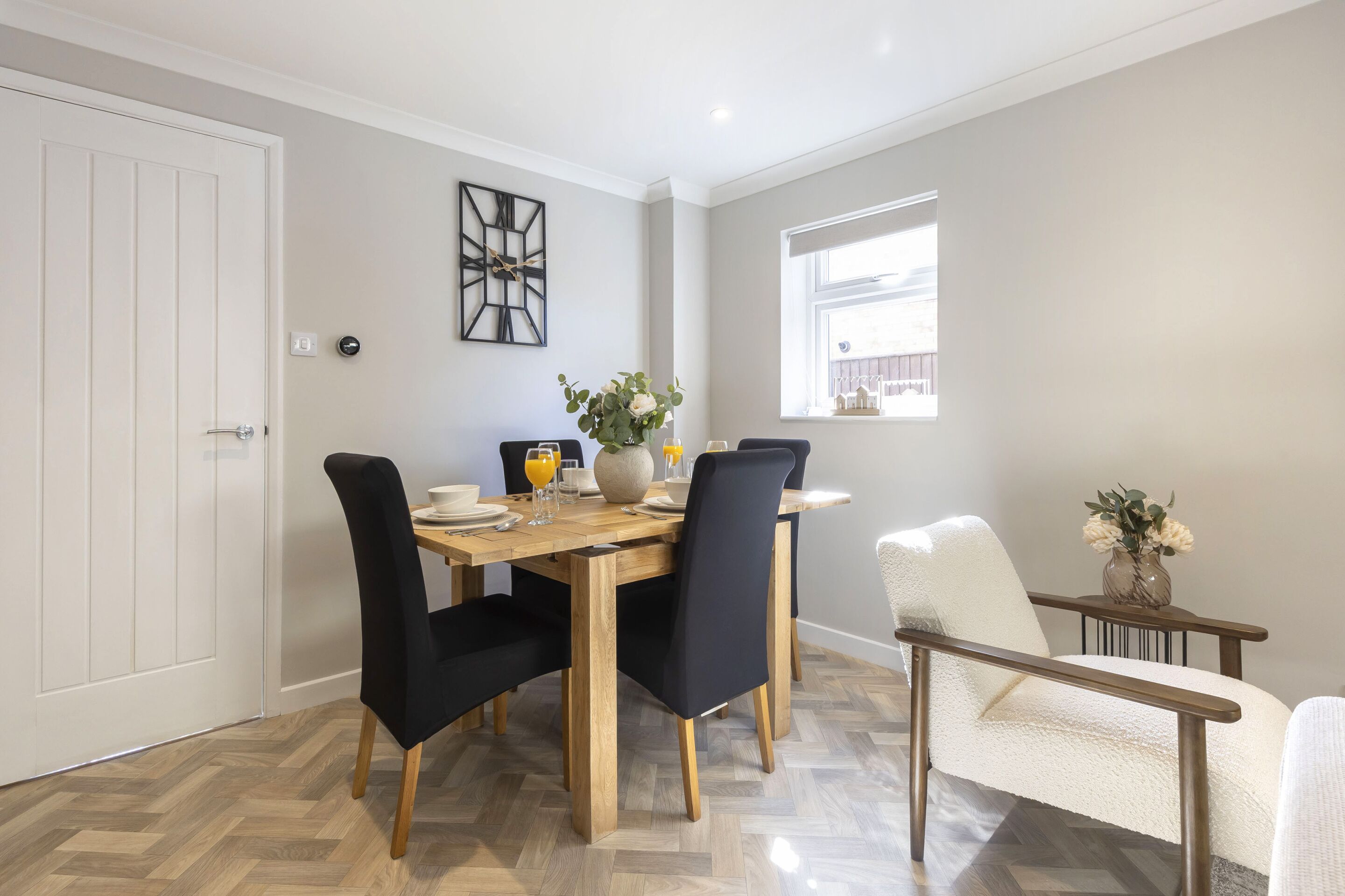 A dining area with a wooden table, black chairs, and a white chair with a cushion. There is a window with a view of a house, and a wall clock.