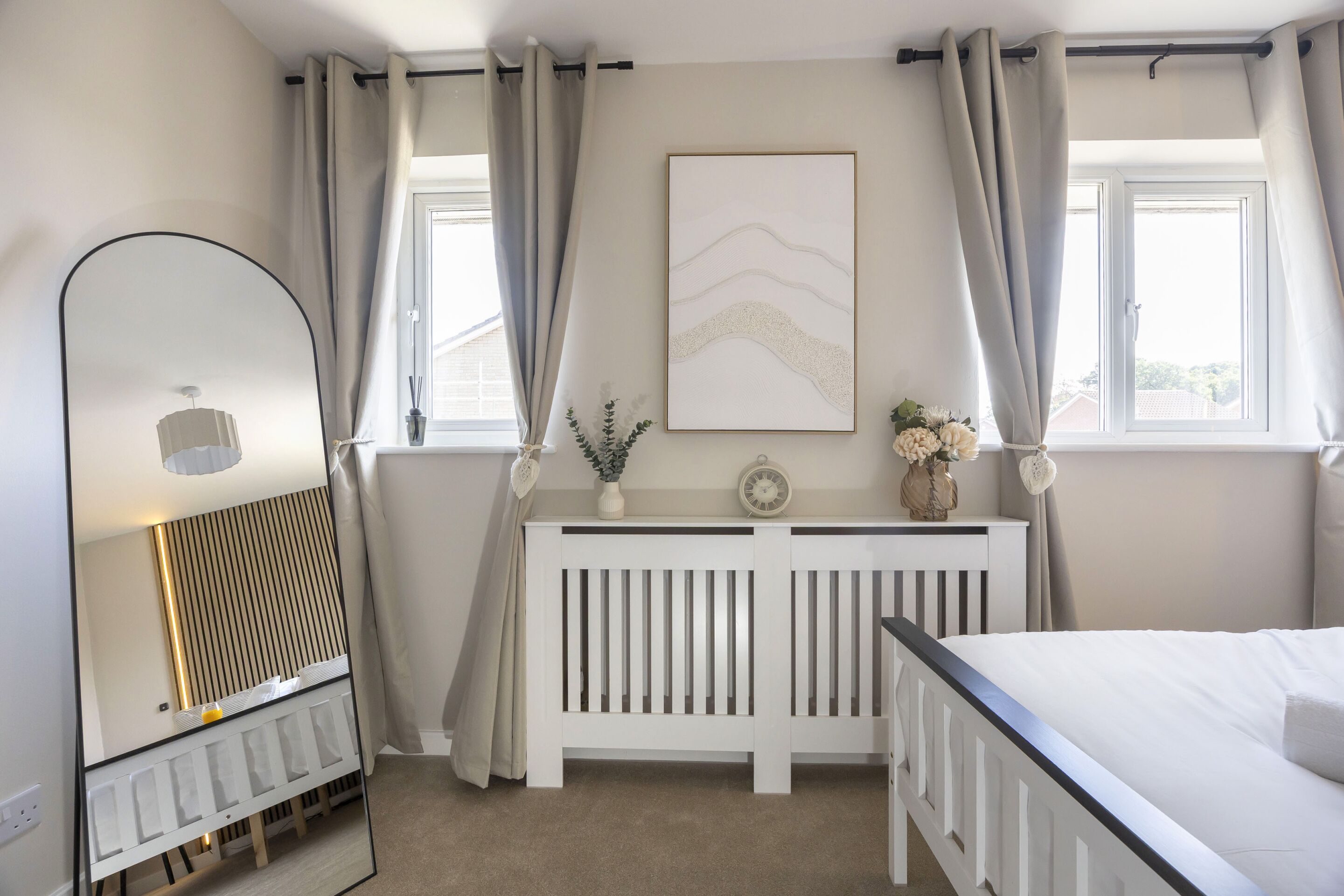 A bedroom with a bed, a bedside table with a clock and flowers, a radiator, and a window with curtains.