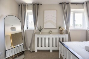 A bedroom with a bed, a bedside table with a clock and flowers, a radiator, and a window with curtains.