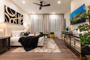 A modern bedroom with a bed, bedside tables, a ceiling fan, a large window with curtains, a TV, and a plant. - 3 Luxury Homes • 12BR • 3 Rooftops • Sleeps 30 (Houston)