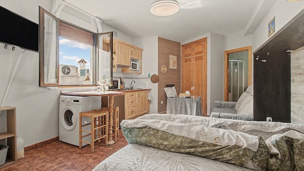 A compact bedroom with a washing machine, a small kitchen area, a bed, and a bathroom.