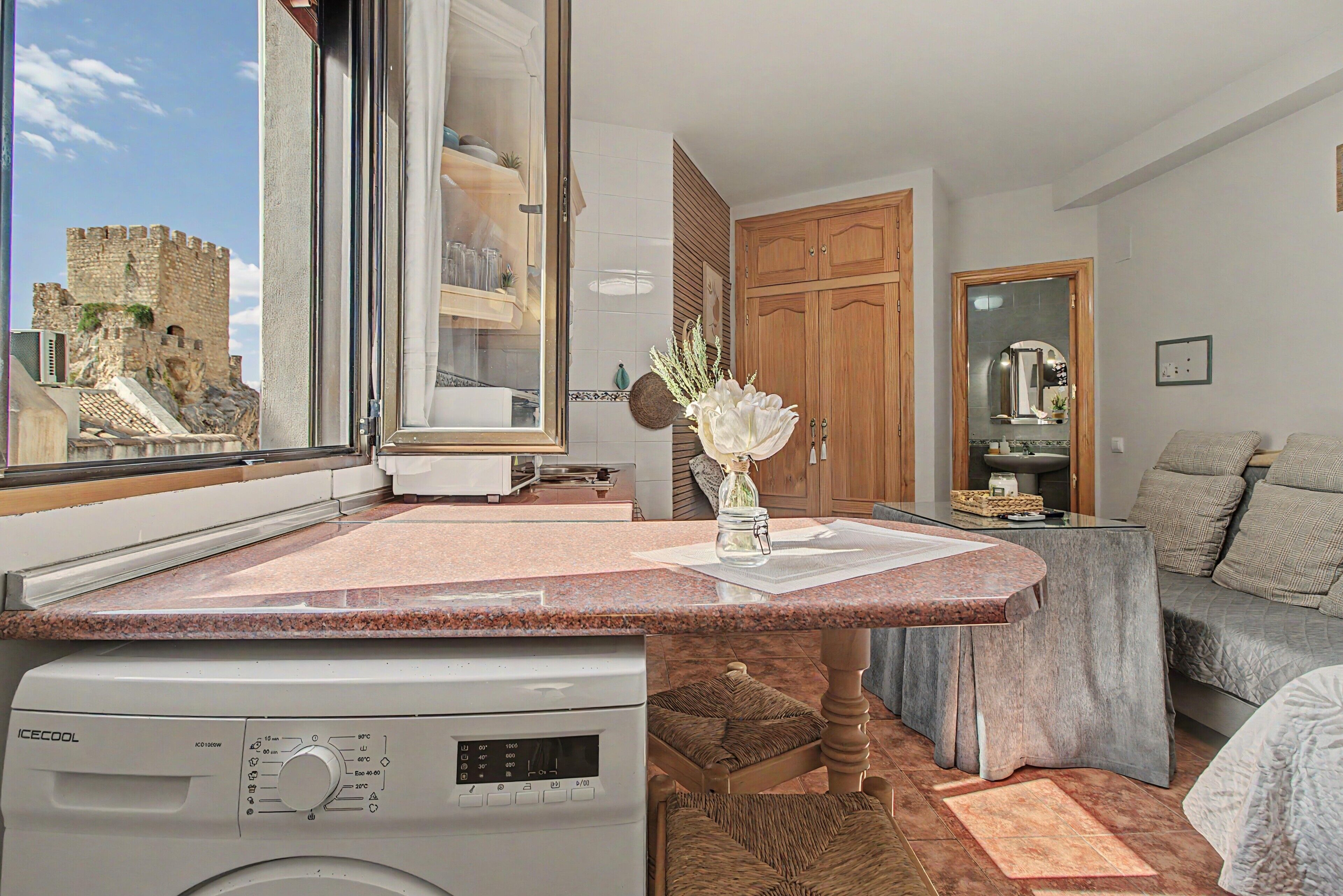 Une cuisine moderne équipée d’un lave-linge intégré, avec vue sur un château par une grande fenêtre, et un coin salon confortable avec un canapé et des coussins.