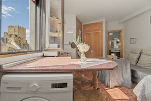 A modern kitchen with a built-in washing machine, a view of a castle through a large window, and a cozy seating area with a sofa and cushions.