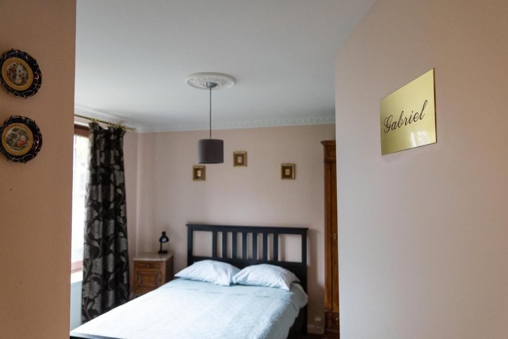 Um quarto com uma cama, uma mesa de cabeceira, um abajur, uma luminária pendente e uma placa na parede com o nome 'Gabriel'.
