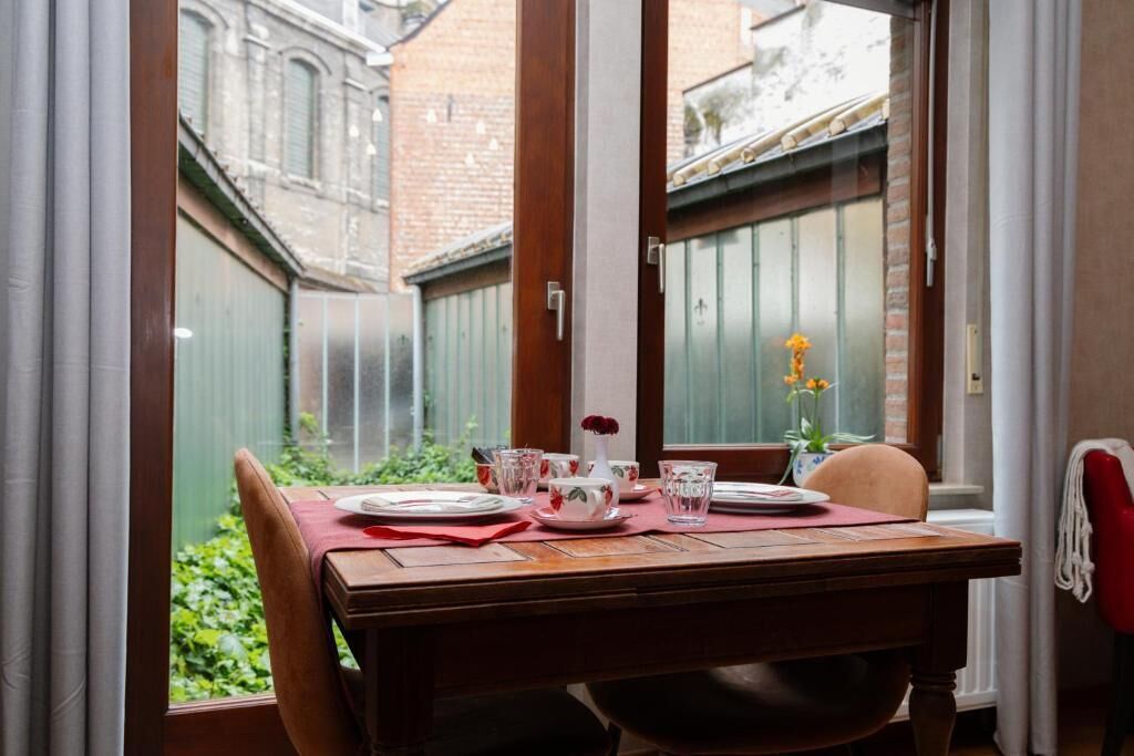 Mesa de jantar posta com pratos, copos e um vaso de flores, posicionada perto de uma janela com vista para um pátio.