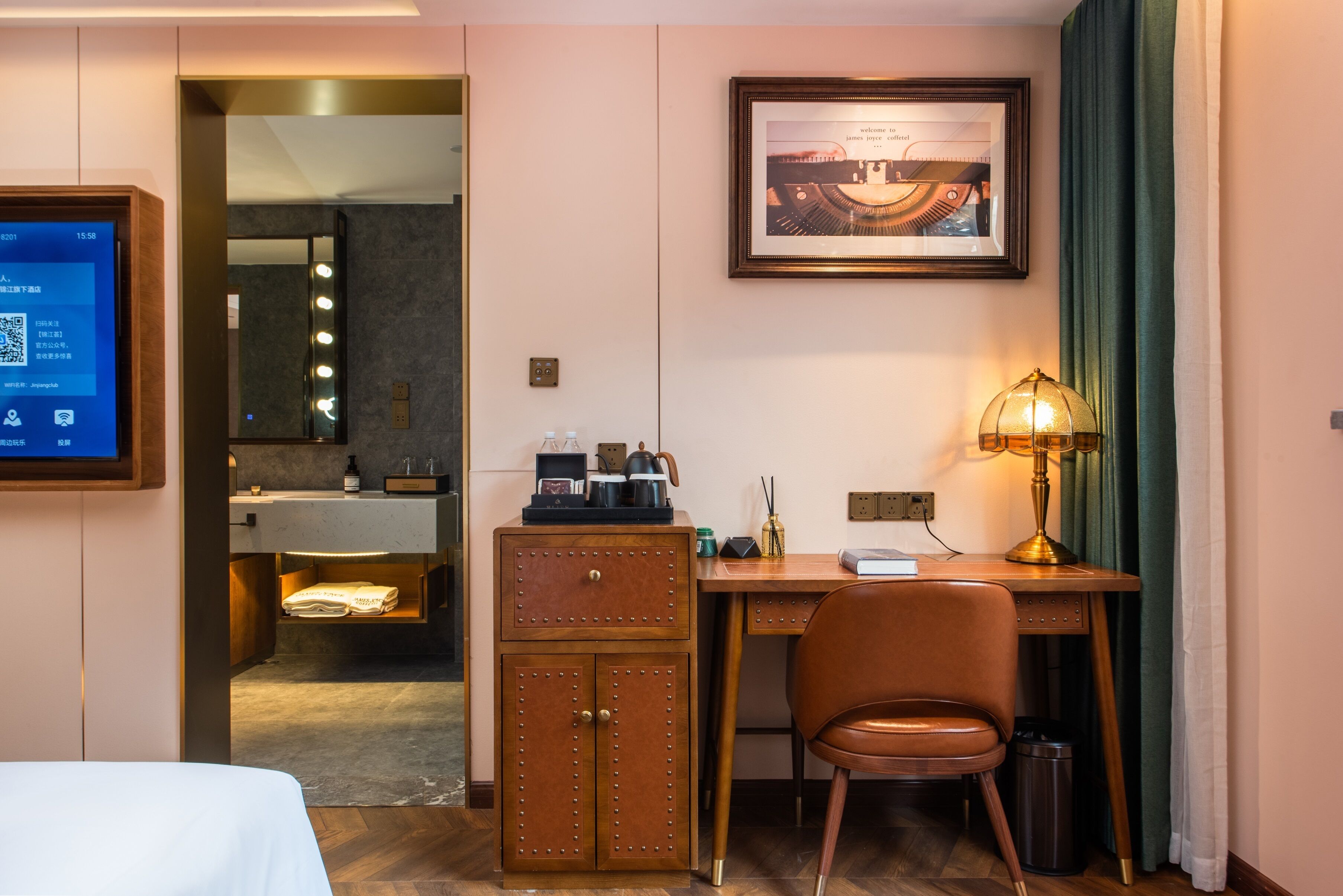 Habitación de hotel con cama, escritorio, silla, lámpara de escritorio, un cuadro en la pared, espejo, tocador con lavabo y un cesto de basura.