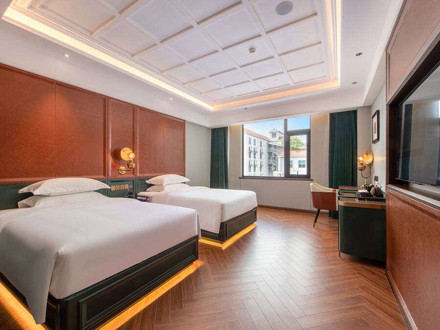 Quarto de hotel com duas camas, uma escrivaninha, uma cadeira, janela com vista para edifícios e teto com iluminação embutida.