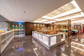 A modern hotel lobby with marble flooring, a central reception desk, and a variety of amenities including a buffet area and a seating area with tables and chairs.